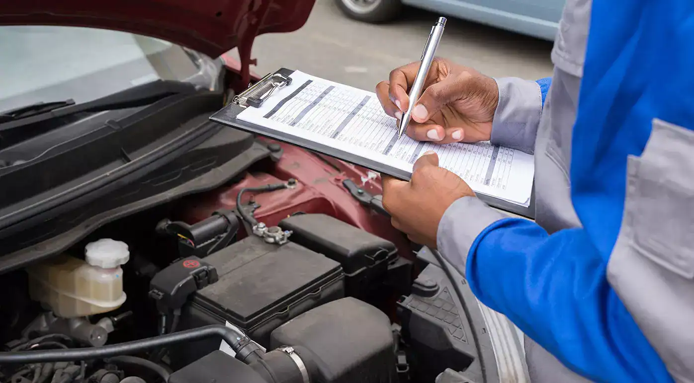 Une personne portant un uniforme bleu et gris remplit une liste de contrôle sur un bloc-notes tout en inspectant le moteur d'une voiture dont le capot est ouvert.